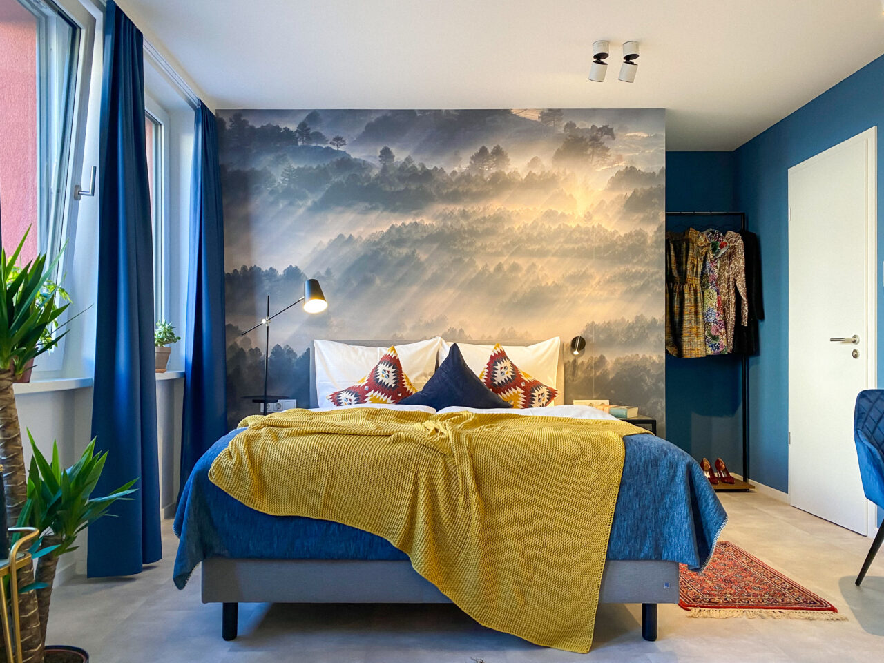 Bett mit senfgelber Decke vor einem Wandbild mit Nebellandschaft